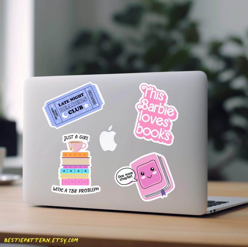 May include: A laptop covered in stickers with a reading theme. Stickers include a blue ticket, a pink book with a face, and text that reads "This Barbie loves books" and "Just a girl with a TBR problem."