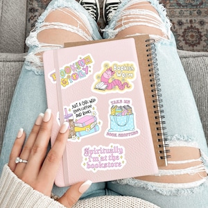 以下が含まれることがあります： 「Fookish Girly」、「Bookish Worm」、「Just a girl who loves coffee and books」、「Take me book shopping」、「Spiritually I'm at the bookstore」と書かれたステッカーが付いたピンク色のノート。 スパイラルノートと破れたジーンズも写っています。