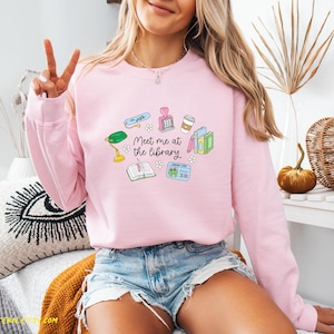 May include: A light pink sweatshirt with a graphic that says "Meet me at the library." The graphic includes a green desk lamp, books, a library card, and other library-themed illustrations. The person is wearing denim shorts.
