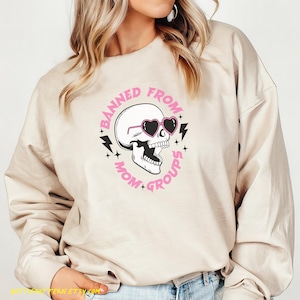 May include: A beige sweatshirt with a pink and black graphic design of a skull wearing heart-shaped sunglasses. The text around the skull reads "Banned from Mom Groups".