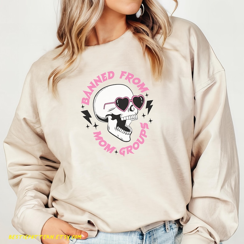 May include: A beige sweatshirt with a pink and black graphic design of a skull wearing heart-shaped sunglasses. The text around the skull reads "Banned from Mom Groups".