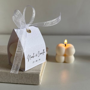 May include: A white favour box with a white satin ribbon and a tag that says "Noah & Camilla 27.08.24". The box is sitting on a beige surface next to a white candle with a flame.