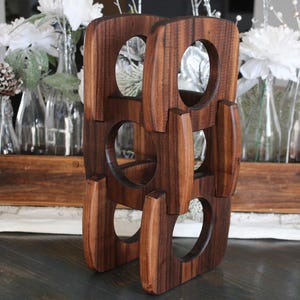 May include: A dark brown wooden wine rack with four openings for bottles. The rack has a unique design with rounded edges and a vertical orientation. The background includes decorative bottles and floral arrangements.