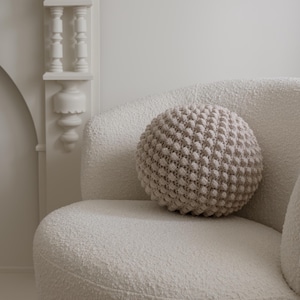 Op de afbeelding: Een witte, pluche fauteuil met een rond, beige gebreid kussen. De stoel staat in een kamer met witte muren en een witte open haard.