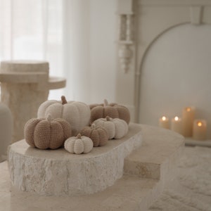 Könnte beinhalten: Eine Sammlung von fünf gehäkelten Kürbissen in verschiedenen Größen, von beige bis braun, auf einer hellbeigen Steinoberfläche angeordnet. Die Kürbisse werden vor einem weißen Kamin mit brennenden Kerzen ausgestellt.