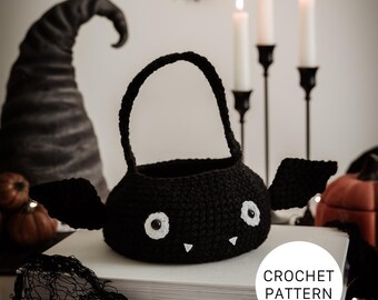 Può includere: Un cestino nero a forma di pipistrello all'uncinetto con occhi e zanne bianche e ali attaccate. Il cestino ha un manico e poggia su una pila di libri. L'immagine include un cappello da strega, candele e pipistrelli decorativi, con il testo "CROCHET PATTERN".