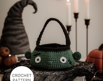 Op de afbeelding: Een groen en zwart gehaakt Frankenstein-thema mandje met witte ogen en oordetails. De mand heeft een zwart handvat en staat op een zwarte kanten doek. De tekst "CROCHET PATTERN" staat in een witte cirkel.