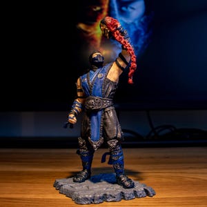 Może przedstawiać: Niebieska i czarna figurka akcji postaci trzymającej czerwoną głowę w dłoni. Figurka stoi na szarej podstawie o skalistej fakturze.