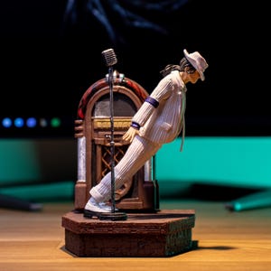 May include: A white figurine of a man wearing a white suit and a white hat, leaning forward with his right arm extended. The figurine is standing on a brown stage with a microphone stand and a red and gold jukebox behind it.