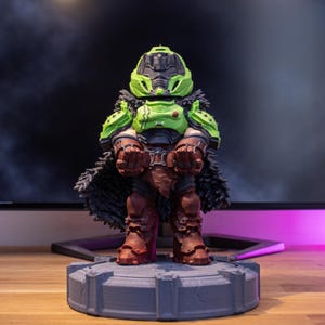 May include: A detailed figurine of a character in green and brown armour, with a black helmet and a fur-like cape. The figure is standing on a grey, circular base. The background is blurred, with a monitor visible.