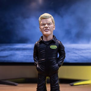 May include: A bobblehead figure of a man with fair hair, dressed in a black jumpsuit with a green and yellow patch. The figure has its hands in its pockets and stands on a wooden surface.