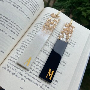 Pode incluir: Dois marcadores de página de resina preta e dourada com detalhes em folha de ouro. Os marcadores de página têm a forma de um retângulo com a parte superior arredondada. Os marcadores de página têm as letras "A" e "M" em dourado na parte da frente.