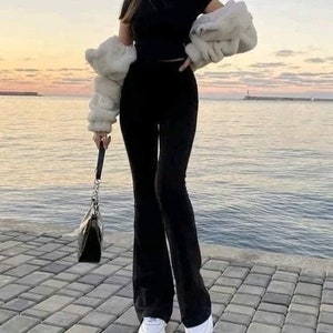 May include: A person wearing a black crop top, flared black trousers, and white trainers. A cream-coloured faux fur jacket is draped over the shoulders. A black handbag with a silver chain is held. The background shows water and a sunset.