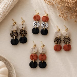 May include: Four pairs of drop earrings featuring three stacked pumpkin-shaped beads each. The beads come in various colors including solid orange, black, cream, and leopard print patterns. Each earring is attached to a gold-toned metal post.
