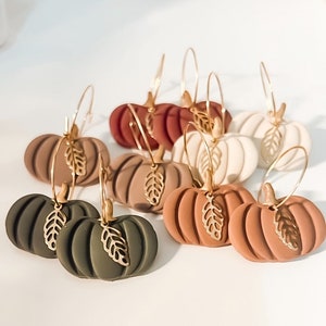 May include: A collection of pumpkin-shaped earrings in various fall colors, including green, brown, and orange. Each earring features a gold-colored leaf charm and a gold hoop. The earrings are designed for a seasonal or autumn theme.