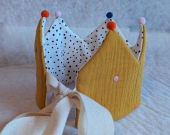 May include: A mustard yellow fabric crown with a white polka dot lining. The crown is decorated with small, colourful pom-poms and a cream-coloured ribbon tie. The crown is designed for a child.