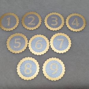 Könnte beinhalten: Ein Set aus neun kreisförmigen, gewellten Papierausstanzungen mit den Zahlen eins bis neun. Jedes Teil hat eine hellblaue, texturierte Mitte mit einer goldglitzernden Zahl und einem goldenen Rand. Die Zahlen sind in drei Reihen angeordnet.