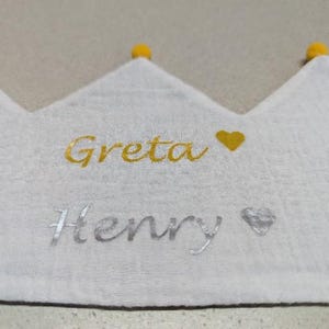 May include: White crown-shaped fabric with the names "Greta" and "Henry" embroidered in gold and silver. A gold heart and silver heart are next to the names. The crown has three points, each topped with a yellow pom-pom.