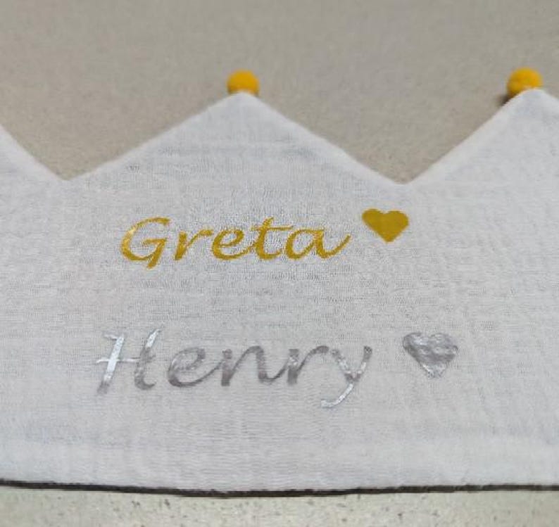 May include: White crown-shaped fabric with the names "Greta" and "Henry" embroidered in gold and silver. A gold heart and silver heart are next to the names. The crown has three points, each topped with a yellow pom-pom.