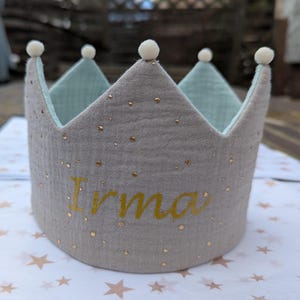 Könnte beinhalten: Eine graue Stoffkrone mit goldenem Glitzer und dem Namen "Irma" in Gold gestickt. Die Krone hat weiße Bommeln oben.