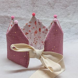 Könnte beinhalten: Eine handgefertigte Stoffkrone in Mauve- und Cremetönen, verziert mit goldenen Sprenkeln und rosa Pompons. Die Innenseite der Krone hat ein Blumenmuster. Ein cremefarbenes Band ist zum Binden befestigt.