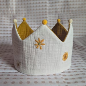 Pode incluir: Uma coroa de tecido branco com uma superfície de musselina texturizada e motivos florais amarelos bordados. A coroa tem um forro interior amarelo mostarda e é rematada com pequenos pompons redondos em cada uma das suas cinco pontas.