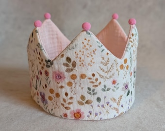 May include: A white crown with a floral pattern and pink pom-poms on the top. The crown is made of fabric and has a pink lining.