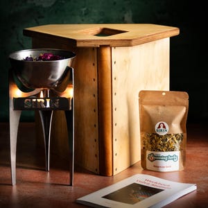 May include: A wooden vaginal steam seat and a stainless steel steaming bowl with the word "SIRIN" on the front. A brown paper bag of herbs and a workbook titled "Workbook Vulva Steam" are also in the image.