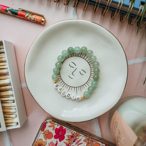 May include: A green stone beaded bracelet with gold accents and white letter beads spelling "BADDIE". The bracelet is on a white ceramic dish with a sun face design.