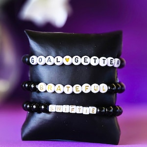 May include: Three black beaded bracelets with white letter beads spelling out "Goal Getter", "Grateful", and "Swiftie". The bracelets are stacked on a black velvet cushion.