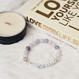 May include: A bracelet made of light purple, gray, and white beads with silver spacers. The bracelet has a charm with the words "Cozy Reader" spelled out in white letters.