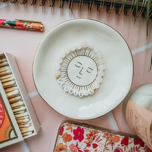 May include: A white ceramic dish with a sun face design holds a beaded bracelet spelling "FEARLESS". A floral phone case, a box of matches, and a pen are also visible. The background is a light pink surface.