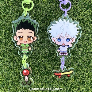 May include: Two acrylic keychains featuring anime-style characters. The left keychain has a green clasp, a character with spiky black hair in a green outfit, and a red ball charm. The right keychain has a purple clasp, a white-haired character, and a skateboard charm.