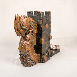 Puede incluir: Una torre decorativa hecha de pequeñas piezas de madera texturizada, pintada en tonos marrones y cobre. La torre tiene una figura de dragón que la envuelve, con un pequeño puente que conduce a la cima.