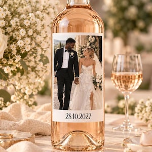 May include: A clear glass bottle filled with rose wine featuring a custom rectangular label. The label displays a photograph of a couple in wedding attire and the date 28.10.2027 printed in black serif font at the bottom of the label.