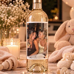 May include: A clear glass wine bottle featuring a rectangular custom photo label on the front. The label displays a colour photograph of two individuals smiling while holding wine glasses. The bottle has a gold-coloured cap and contains clear liquid.
