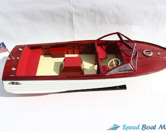 Può includere: Un modello di motoscafo con ponte in legno rosso e scafo bianco. L'interno presenta sedili rossi e un pavimento color crema. Una bandiera americana è attaccata a poppa. La barca ha un volante e un parabrezza.