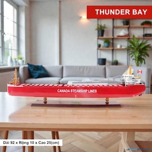 Pode incluir: Modelo de navio vermelho com o texto "CANADA STEAMSHIP LINES" na lateral. O modelo é montado em uma base de madeira. O texto "THUNDER BAY" é exibido em uma faixa vermelha. O navio mede aproximadamente 92 cm de comprimento, 10 cm de largura e 25 cm de altura.