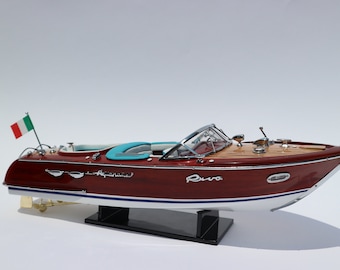 May include: A wooden model of a Riva Aquarama speedboat with a teal interior, a black stand, and an Italian flag on the bow. The boat is labeled "Riva Aquarama" and "Riva".