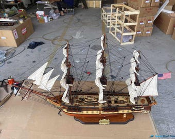 Può includere: Modello in legno dettagliato di una nave alta con vele bianche e una bandiera americana. La nave presenta un'intricata attrezzatura, alberi multipli e uno scafo marrone scuro. Il modello è esposto su una base di legno.