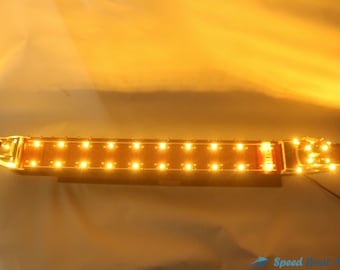 May include: A wooden model ship with a brown hull and a white deck. The ship has a row of yellow lights along the deck and a black cord extending from the back. The ship is on a white surface.