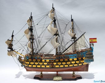 May include: A detailed model of a sailing ship with yellow, blue, and brown accents. The ship has multiple masts with white sails and intricate rigging. A Spanish flag is visible at the stern. The model is mounted on a wooden base.