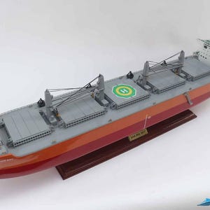 Pode incluir: Um modelo detalhado de um navio de carga vermelho e cinza com um heliporto verde. O navio está sobre uma base de madeira e tem o nome "HALONG BAY" impresso no lado.