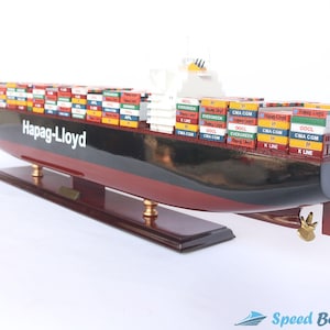 May include: A detailed model of a container ship with a red and black hull. The ship is loaded with colorful shipping containers, many of which have the logos of shipping companies like Hapag-Lloyd, CMA CGM, OOCL, and K Line. The model is on a wooden base.