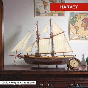 May include: A detailed wooden model sailboat, labeled "HARVEY", with cream-colored sails and intricate rigging. The model is displayed on a dark wooden surface. The dimensions are 23.6 inches long, 7.1 inches wide, and 26.8 inches high.