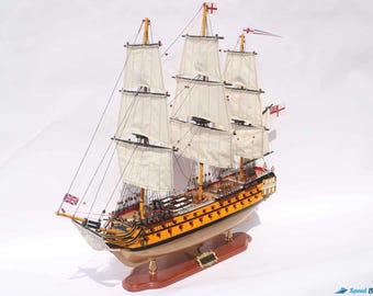 Può includere: Modello in legno dettagliato di una nave alta con più alberi e vele. La nave presenta uno scafo giallo, vele bianche e un complesso sartiame. Sono visibili bandiere con la Union Jack e altri emblemi. Il modello è montato su una base di legno.
