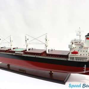 Pode incluir: Modelo detalhado de um navio de carga, com casco vermelho, linha d'água preta e superestrutura branca. O modelo inclui guindastes, um heliporto e uma hélice. O navio é montado em uma base de madeira escura. O texto "Speed Boat Model" é visível.