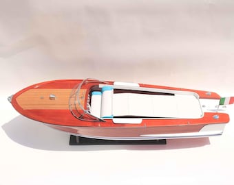 May include: A model wooden speedboat with a red hull and a light wood deck. The boat features white seating, blue accents, and a small Italian flag on the stern. The boat is displayed on a black stand.