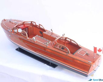 May include: A wooden model of a vintage speedboat with a mahogany finish. The boat has a Canadian flag on the back and is displayed on a black stand.