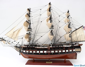 May include: A detailed wooden model of a tall ship with three masts, tan sails, and black rigging. The ship has a dark brown hull and a small American flag at the stern. The model is mounted on a wooden base.
